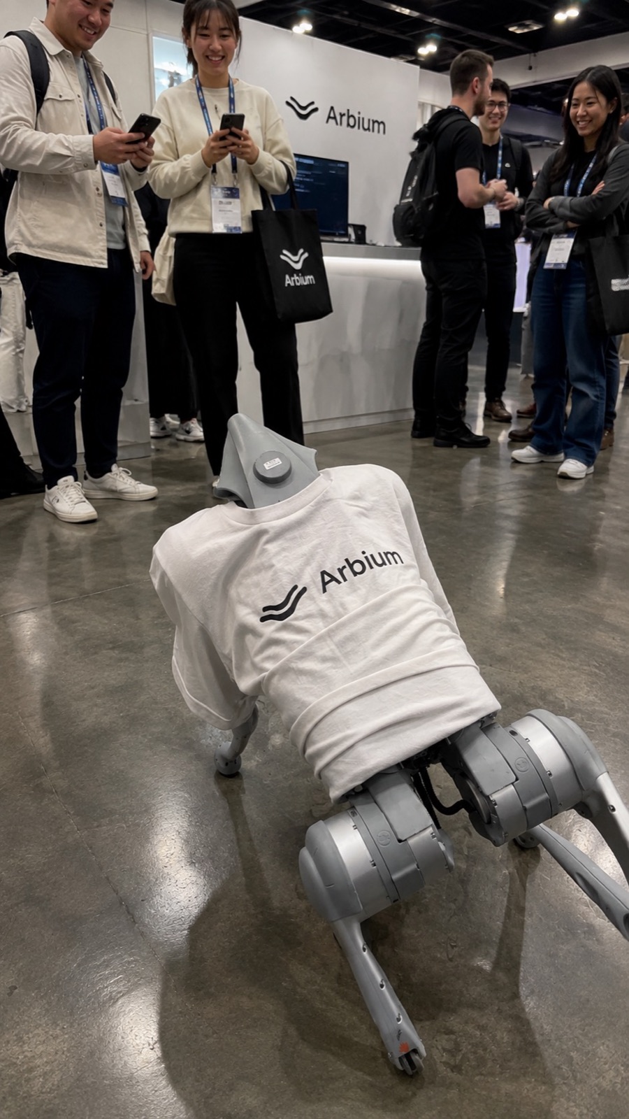 The robot dog in a white Arbium t-shirt on a conference floor, with attendees filming it on their phones
