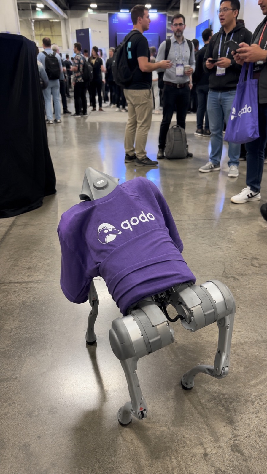 The robot dog in a Qodo t-shirt walking down an aisle between conference booths